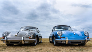 Två klassiska Porsche 356 i grått och blått sida vid sida på en äng